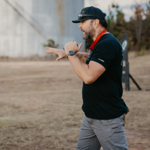 A man wearing a cap, sunglasses, and a smartwatch is outdoors, gesturing with his hands as he speaks about Tactical Carbine Training.