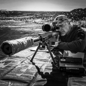 A man is aiming through the scope of a rifle mounted on a bipod in an open, hilly landscape. There is ammunition and a box on the table next to him. The black and white scene captures a moment of tactical carbine training.