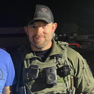 A person wearing a tactical uniform and cap stands outdoors at night, partially smiling. They are equipped with a body camera, radio, and other tactical gear, possibly after completing Tactical Carbine Training.