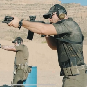 Two individuals engaging in Tactical Carbine Training outdoors, both wearing tactical gear and using rifles. The person in the foreground is aiming while the other in the background is also seen taking aim.