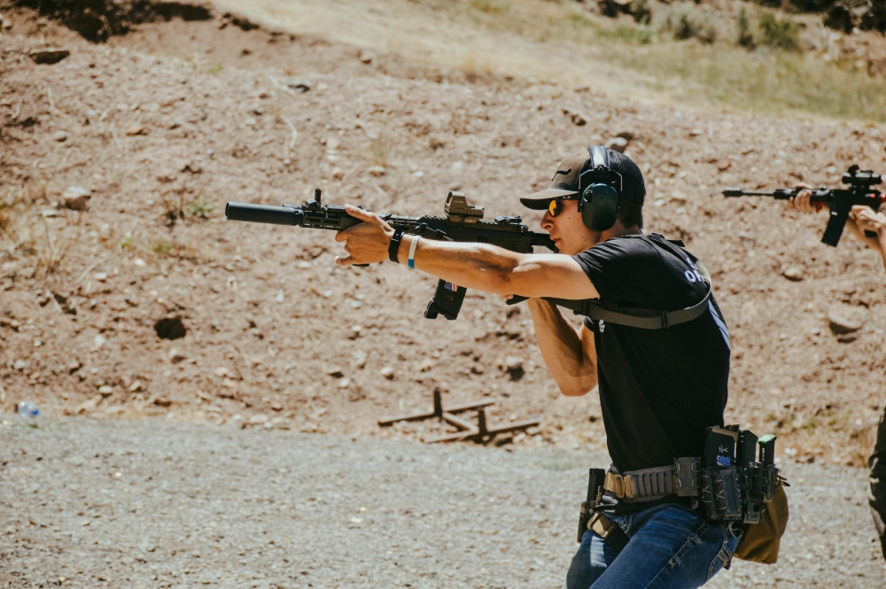 At an outdoor shooting range, a person wearing ear protection and a black shirt focuses intently while aiming a rifle. Another individual in the background also aims their rifle, engaged in tactical carbine training.