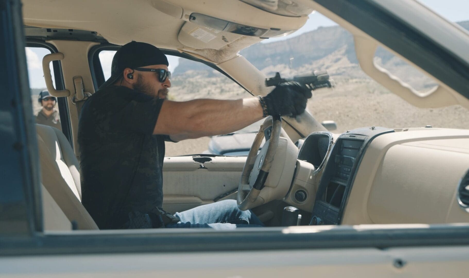 A man in tactical gear, clearly engaged in Tactical Firearms Training, aims a handgun outside the window of a vehicle in a desert setting.