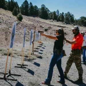 An instructor assists a participant in aiming a firearm during their Private Training (Session Deposit) at the shooting range, with paper targets positioned ahead, as other individuals engage in practice in the background.