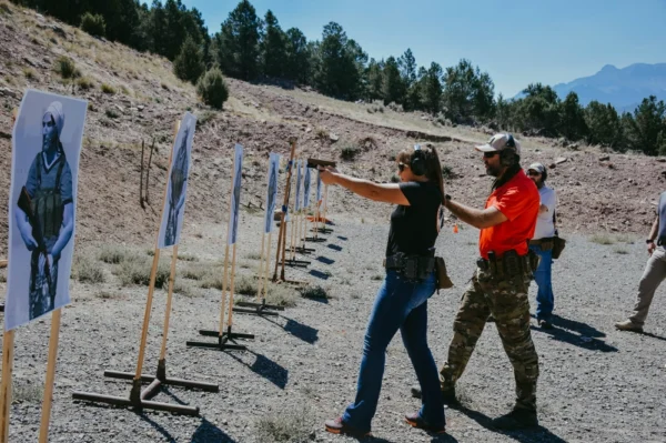 An instructor assists a participant in aiming a firearm during their Private Training (Session Deposit) at the shooting range, with paper targets positioned ahead, as other individuals engage in practice in the background.